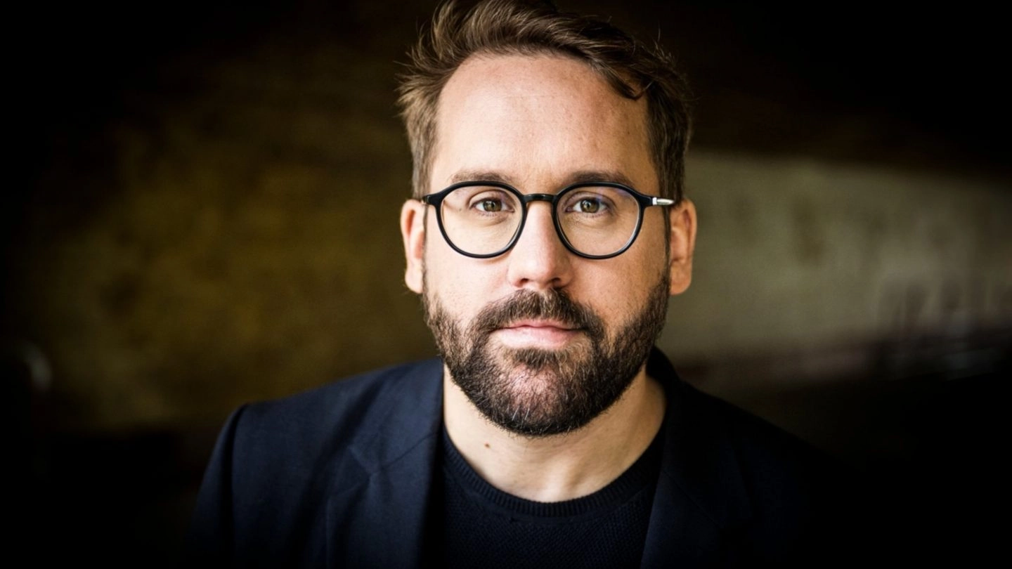 Porträt eines Mannes mit Brille und Bart in dunklem Sakko vor dunklem Hintergrund