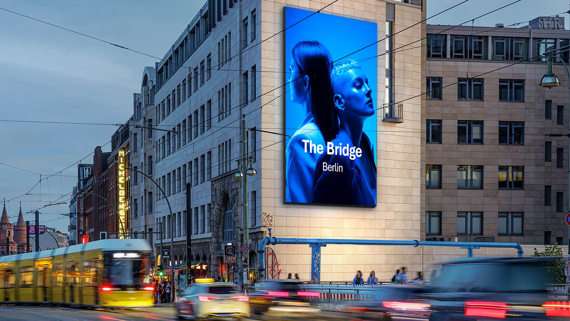 Hochformatiger LED-Screen mit der Aufschrift The Bridge Berlin an einer Hausfassade an der Warschauer Brücke, davor Straßenbahn und Autoverkehr in der Dämmerung