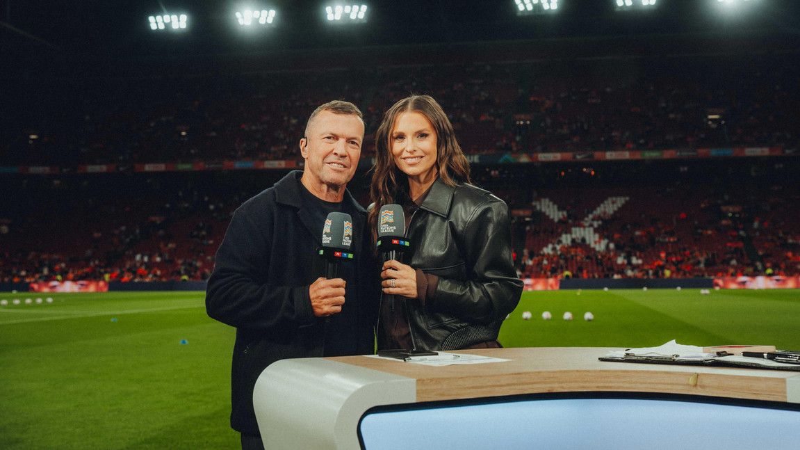 Zwei RTL-Moderatoren mit Mikrofonen am Moderationspult im Fußballstadion