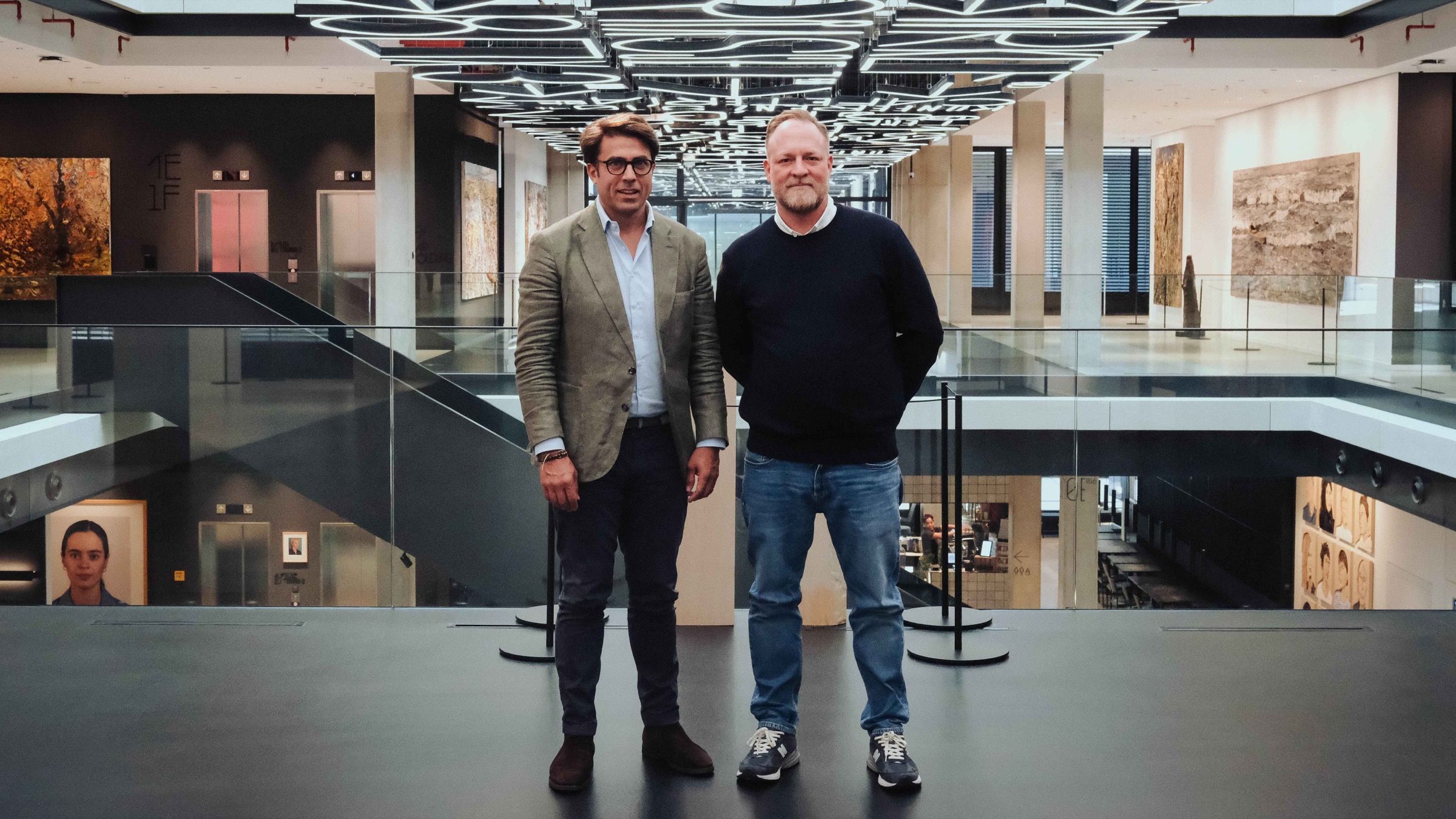 Zwei Männer stehen nebeneinander auf einer Galerie in einem hohen Atrium mit Lichtinstallation an der Decke und großformatigen Gemälden an den Wänden