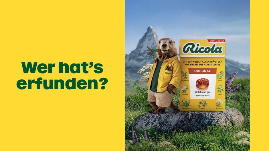 Murmeltier Ric in gelber Jacke lehnt an einer Packung Ricola vor Alpenkulisse, links der Schriftzug „Wer hat's erfunden?“