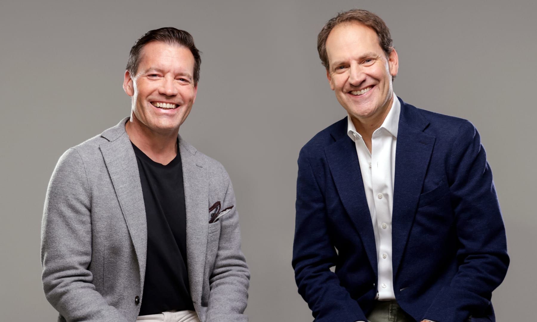 Jon Cook und Eric Campbell lächelnd nebeneinander vor grauem Hintergrund