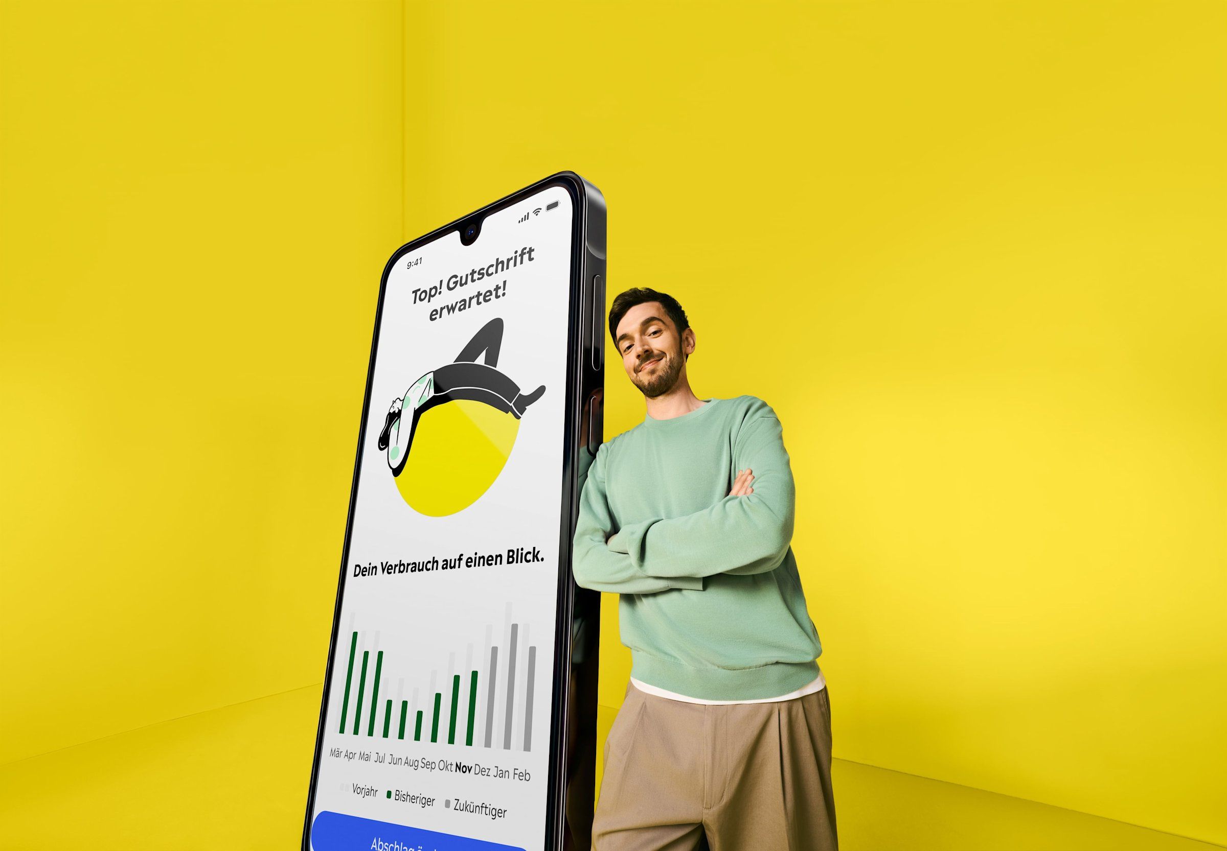 Ein Mann im hellgrünen Pullover lehnt in einem gelben Raum an einem übergroßen Smartphone, auf dem die Yello App den Stromverbrauch zeigt